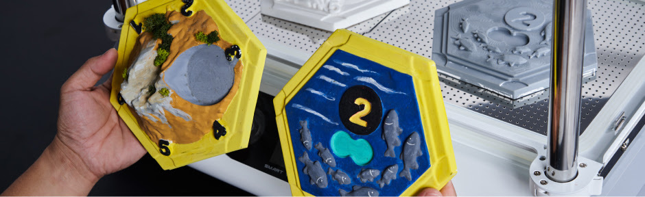 La mano regge due stampi in plastica HIPS dipinti di una tessera esagonale di Catan 3D